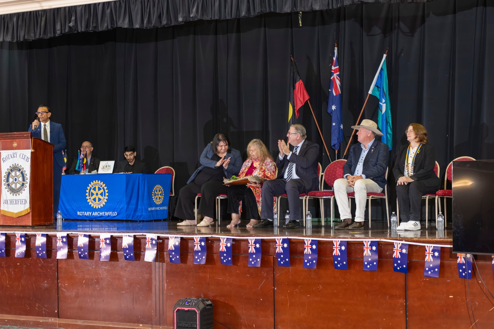 A speaker at the Rotary Club of Archerfield lectern during the Australian Citizenship Ceremony, with guests seated on stage beside the Australian, Aboriginal and Torres Strait Islander flags