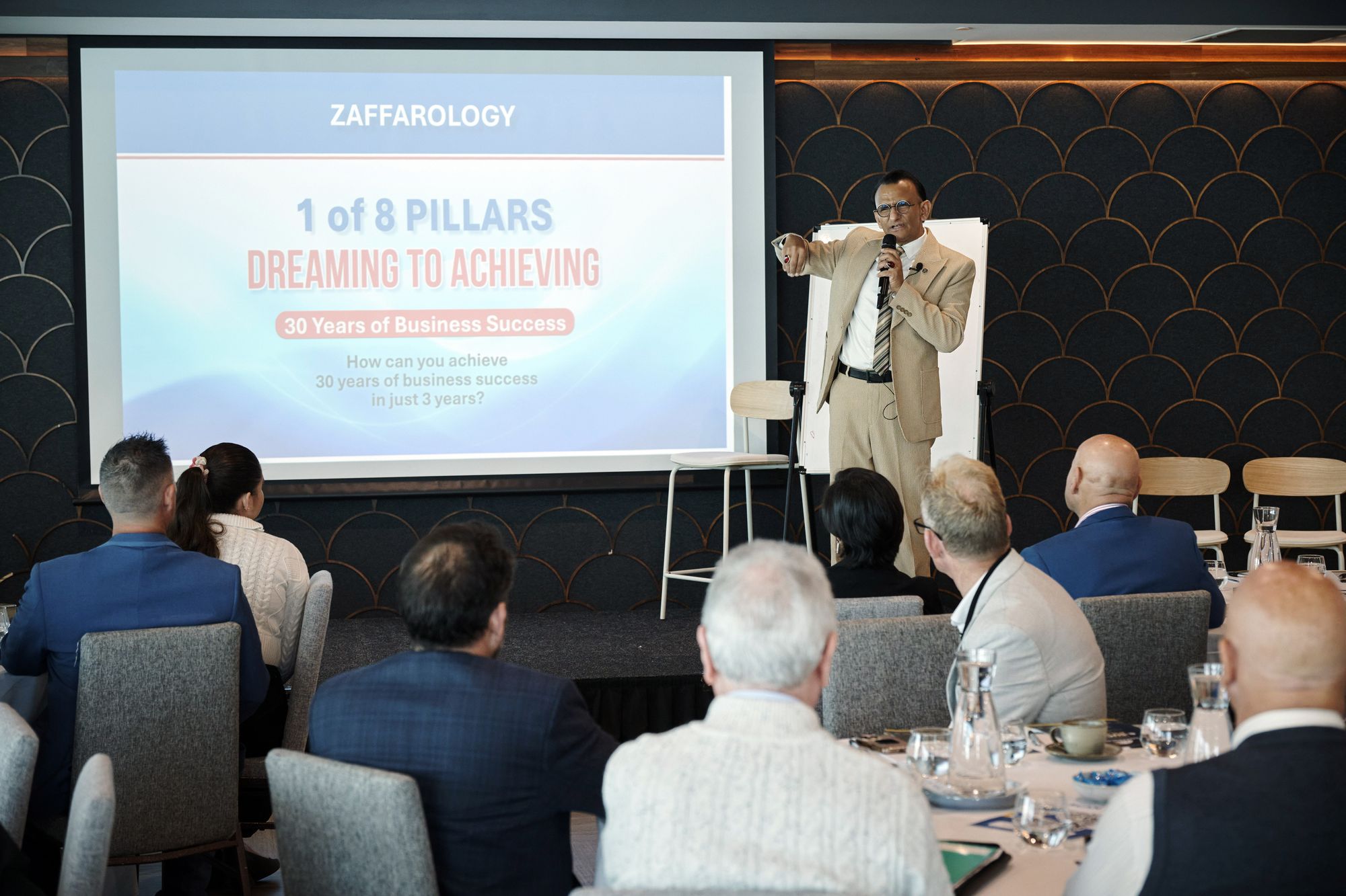Zaffar Khan pointing to the audience beside a slide reading 1 of 8 Pillars, Dreaming to Achieving