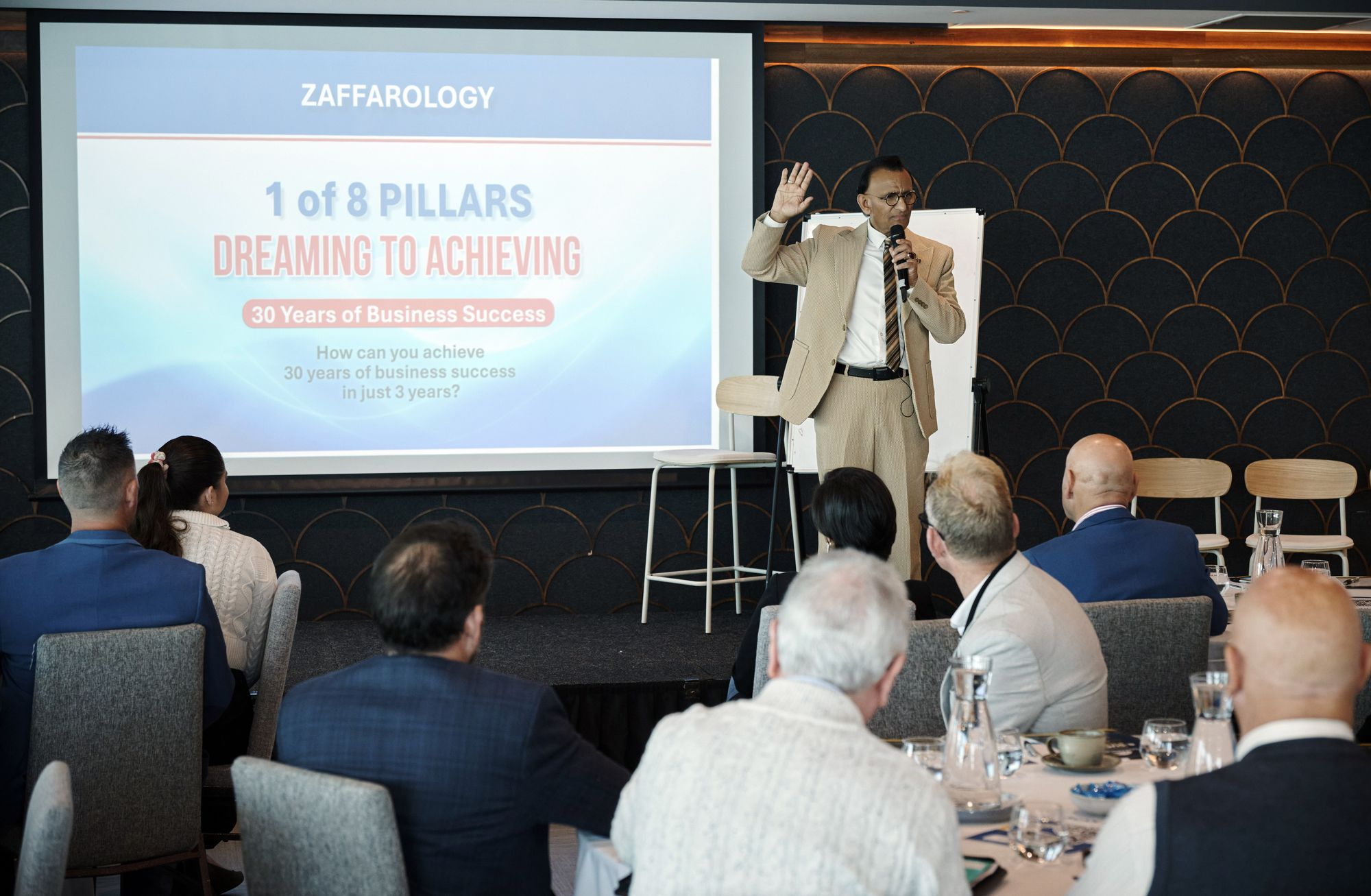 Zaffar Khan with a hand raised mid sentence in front of the 8 Pillars slide