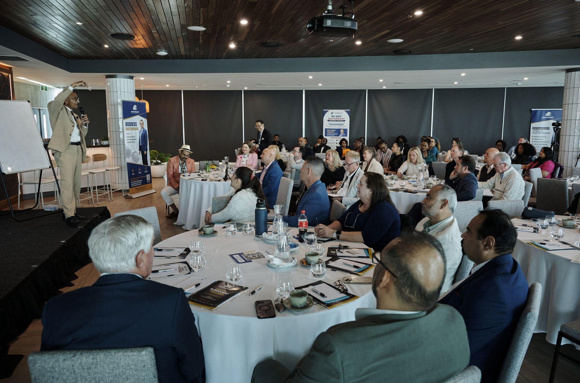 Zaffar Khan addressing the room from the stage beside a Business Matchmaking banner