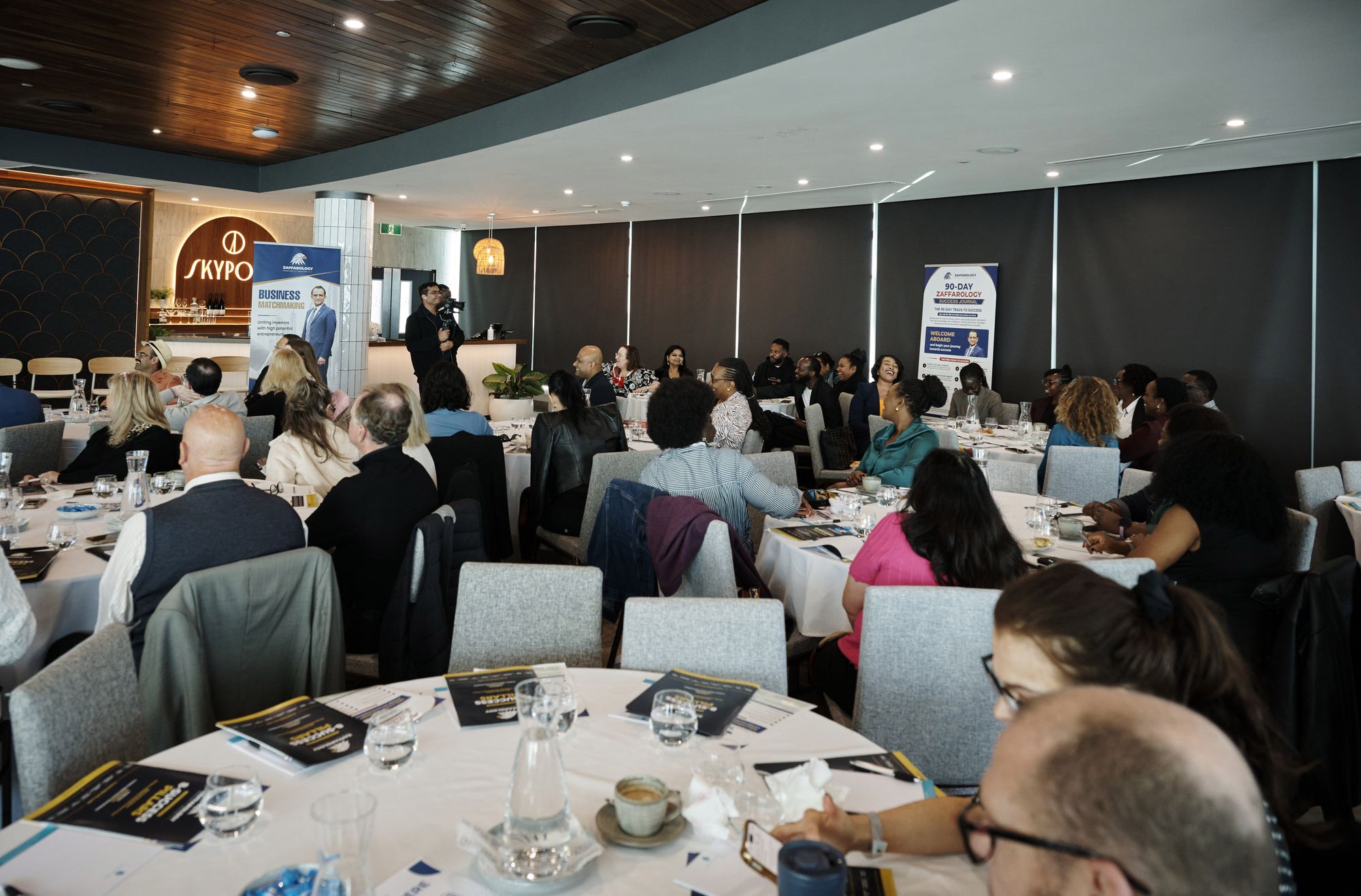 The room mid session with attendees at round tables and the SkyPoint bar behind